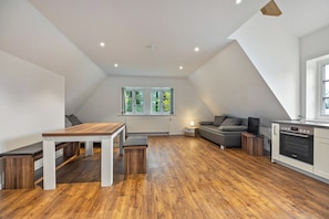 A modern loft apartment with a dining area, seating, a kitchen, and a large window with a view of greenery.