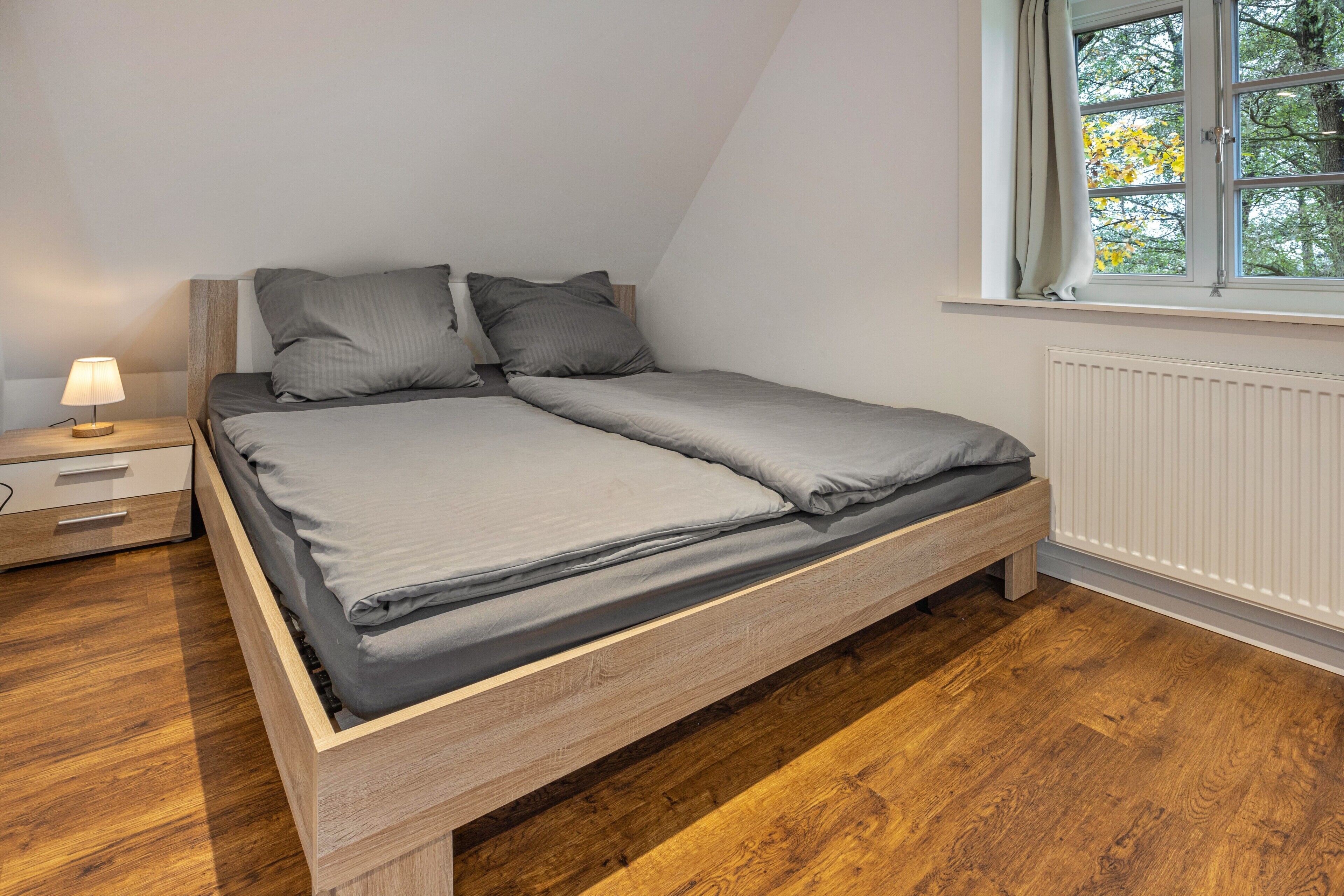 Ein Einzelbett mit Holzrahmen, ein Nachttisch mit Lampe und ein Fenster mit Blick auf Bäume.