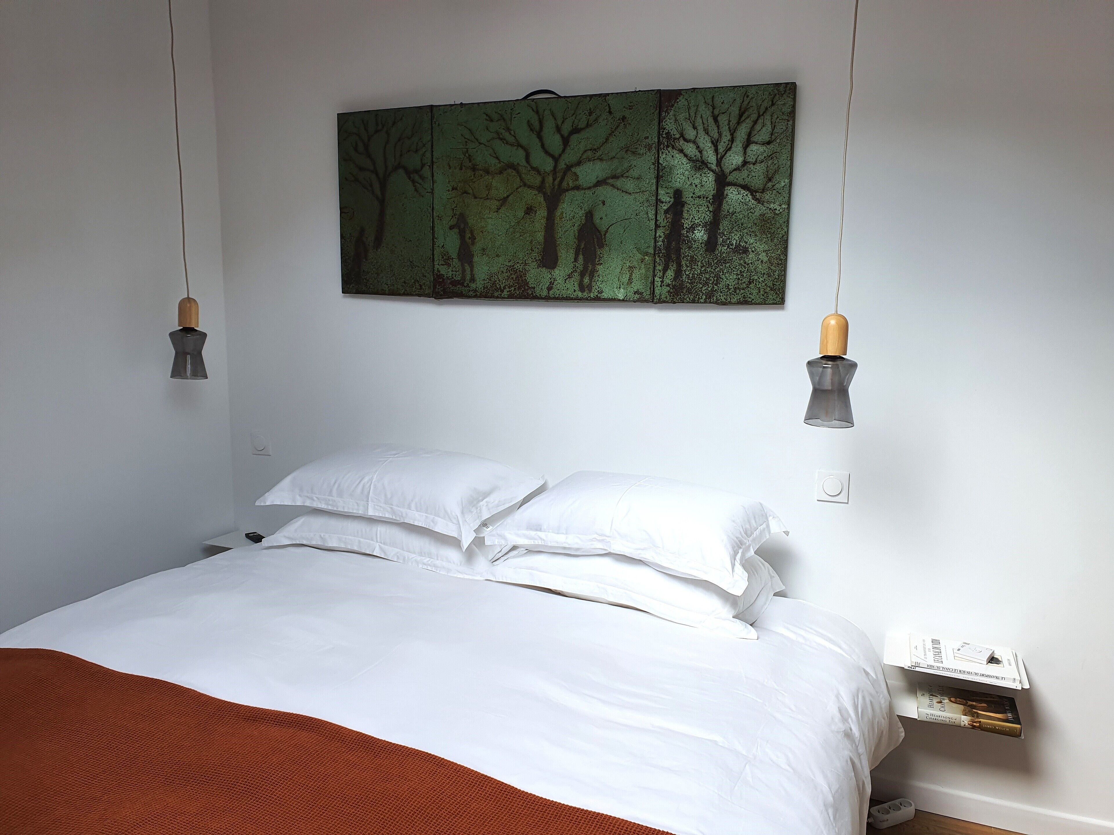 A neatly made bed with white linens and a brown throw blanket, a green wall art piece above the bed, and two wall-mounted lamps.