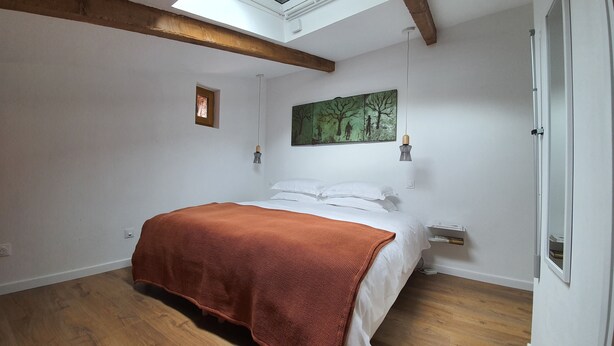 Un dormitorio con tragaluz, vigas de madera, una cama con sábanas blancas y una manta naranja, una mesita de noche y un cuadro en la pared.