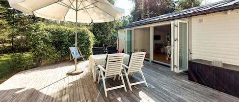 Una terraza al aire libre con mesa y sillas, un paraguas grande y vistas a una casa con la puerta abierta.