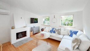 Ein modernes Wohnzimmer mit Kamin, einer Sofagarnitur, einem Fernseher und zwei Fenstern mit Blick auf Grünflächen.