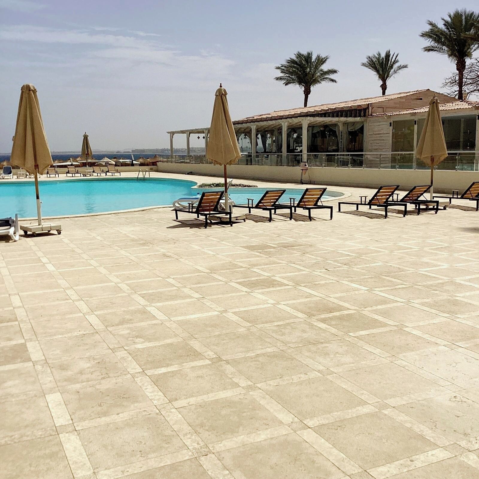 A poolside area with lounge chairs and umbrellas, a clear sky, and a building with a tiled roof.