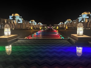Vista nocturna de un patio de hotel con fuente de agua, iluminado con luces de colores, rodeado de arquitectura tradicional con estructuras abovedadas.