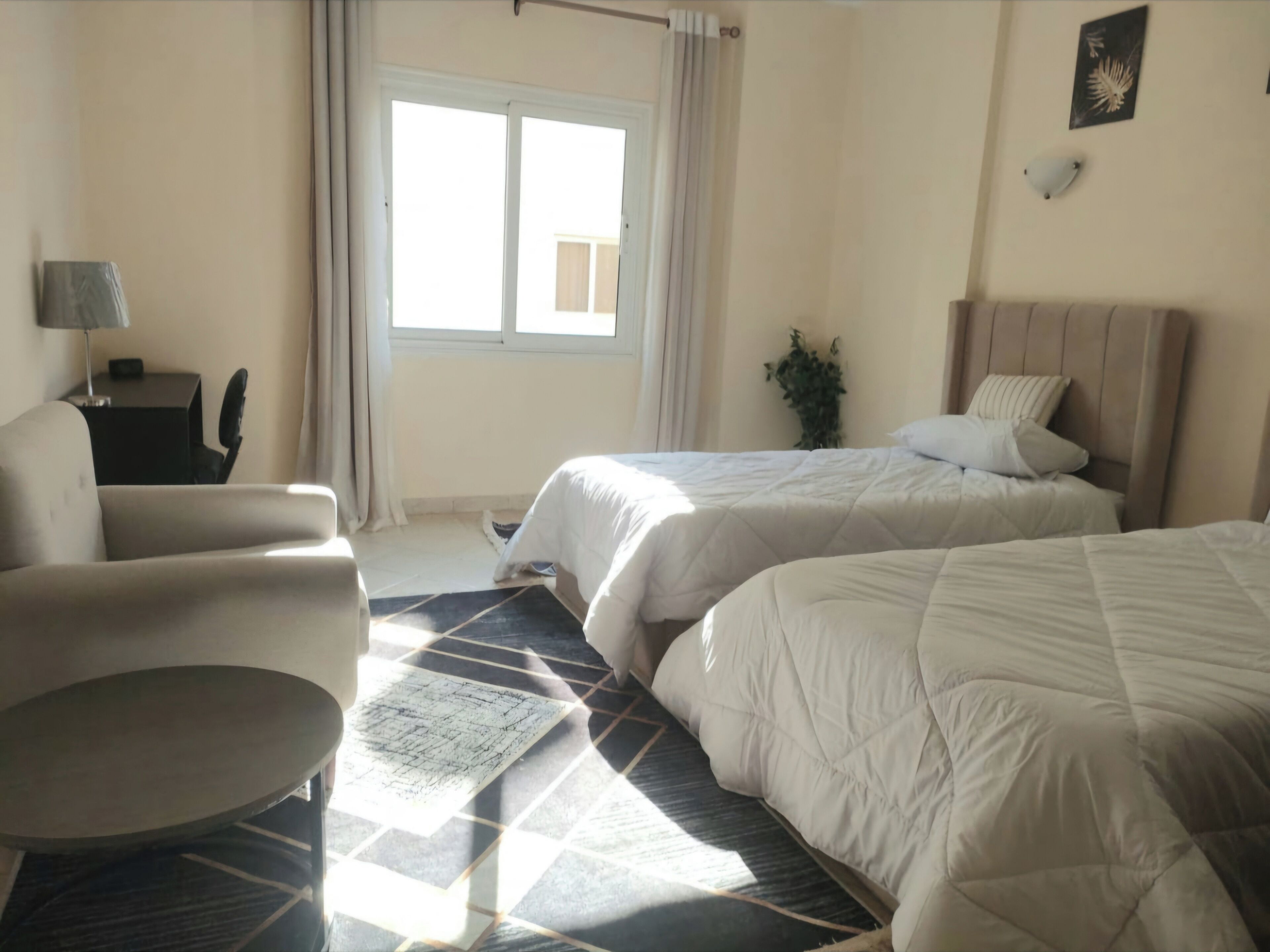 A hotel room with two beds, a desk, a chair, a window with curtains, and a plant.