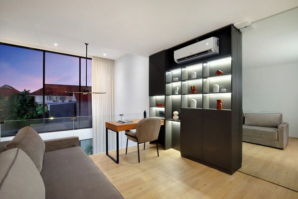 A modern living room with a large window, a sofa, a desk, and a built-in shelving unit with decorative items.