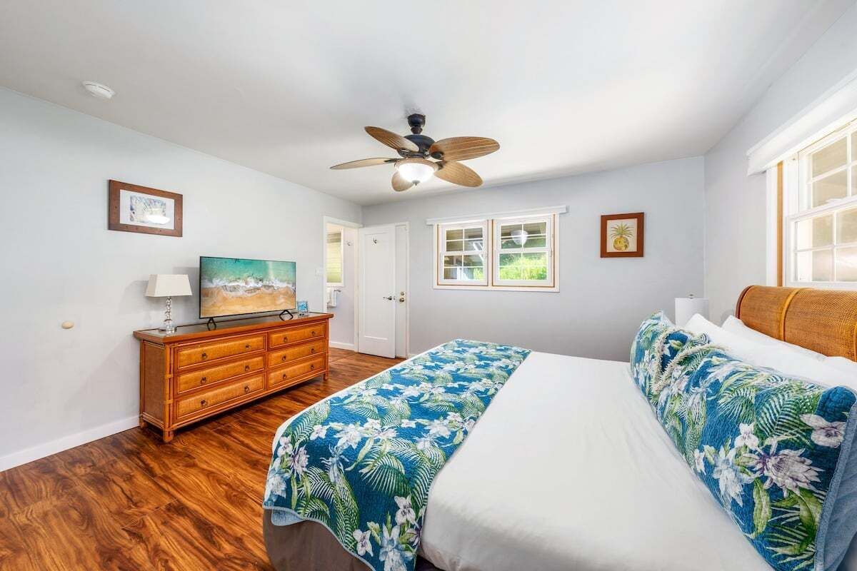 A bedroom with a wooden dresser, a bed with a floral bedspread, a ceiling fan, and a window with a view of greenery.