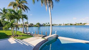 A swimming pool with a curved edge, surrounded by palm trees and a view of a body of water and distant buildings.