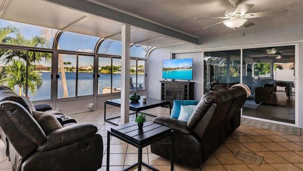 A spacious living area with a large window overlooking a body of water, a ceiling fan, and a flat-screen TV.