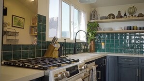 Cucina con piano cottura a gas, forno e lavello. Si vedono mensole con oggetti decorativi e una pianta in vaso.