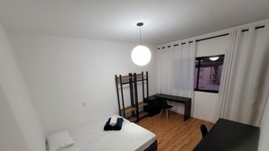 Een kleine kamer met een bed, een bureau en een stoel. Er is een raam met gordijnen, een hanglamp en uitzicht op gebouwen.