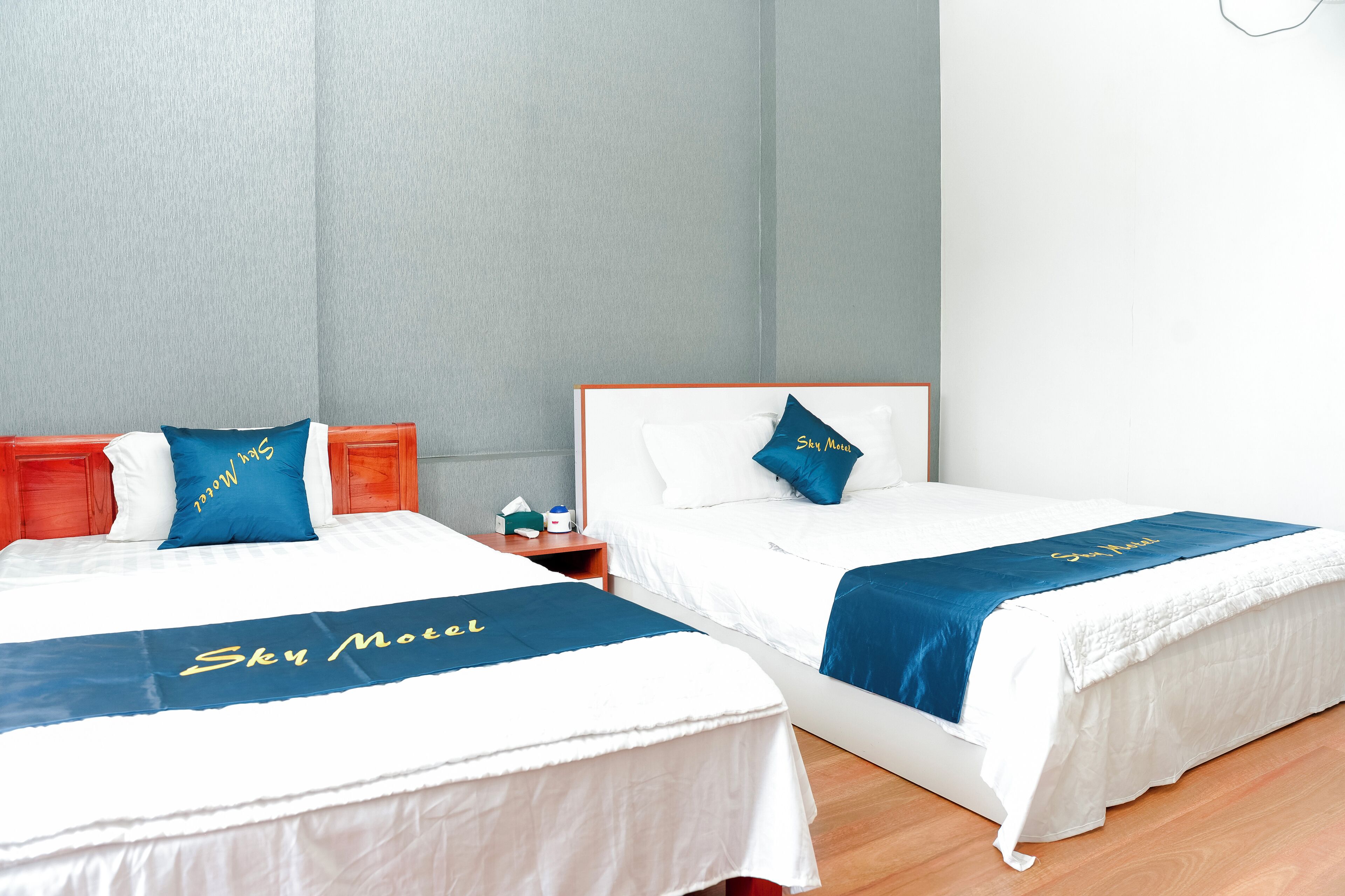 Twee bedden met witte beddengoed en blauwe hoofdborden, elk met een kussen en een blauwe loper met de tekst 'Sky Motel'.