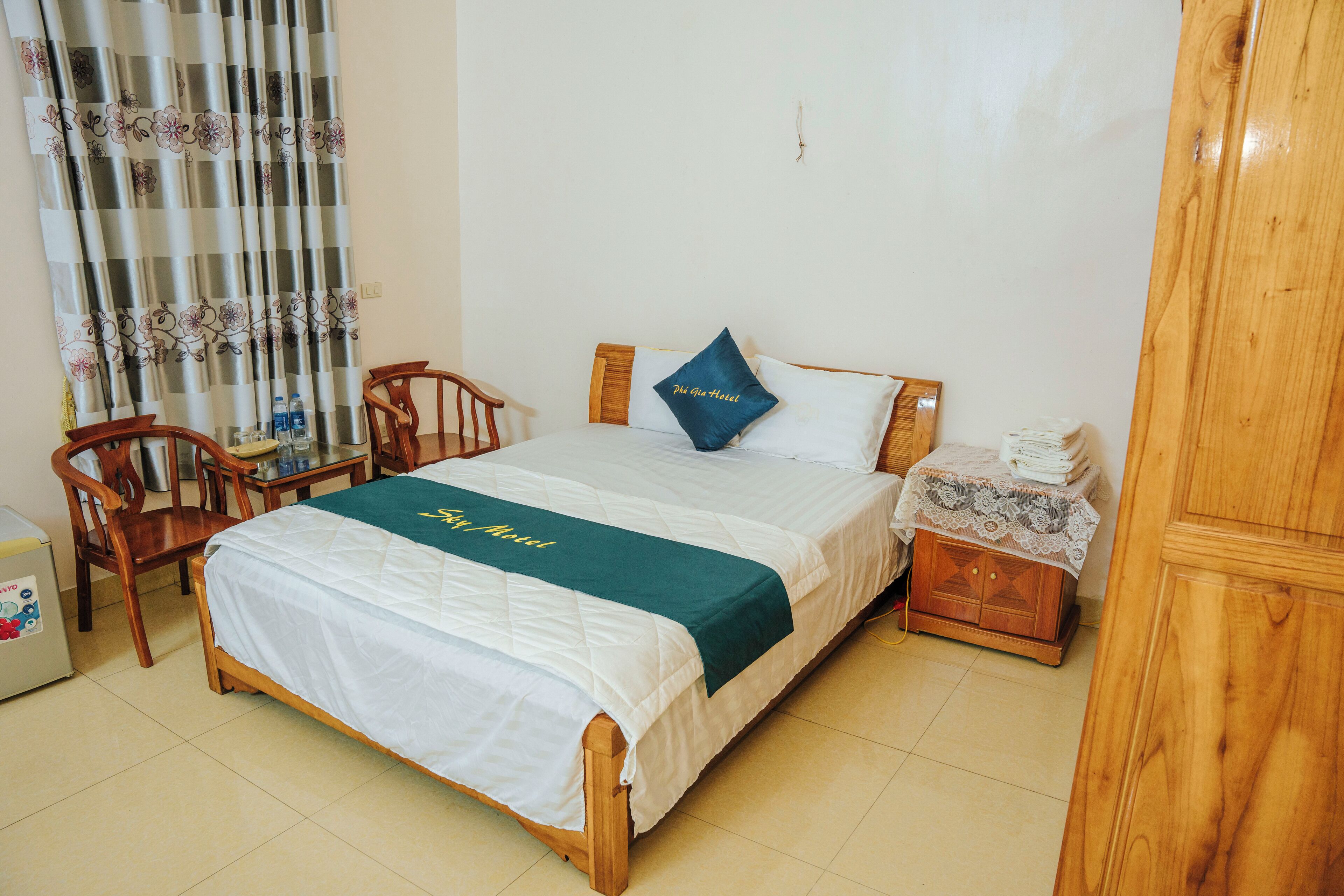 A hotel room with a bed, a wooden nightstand, a chair, a table with a bottle, and a curtain with floral patterns.