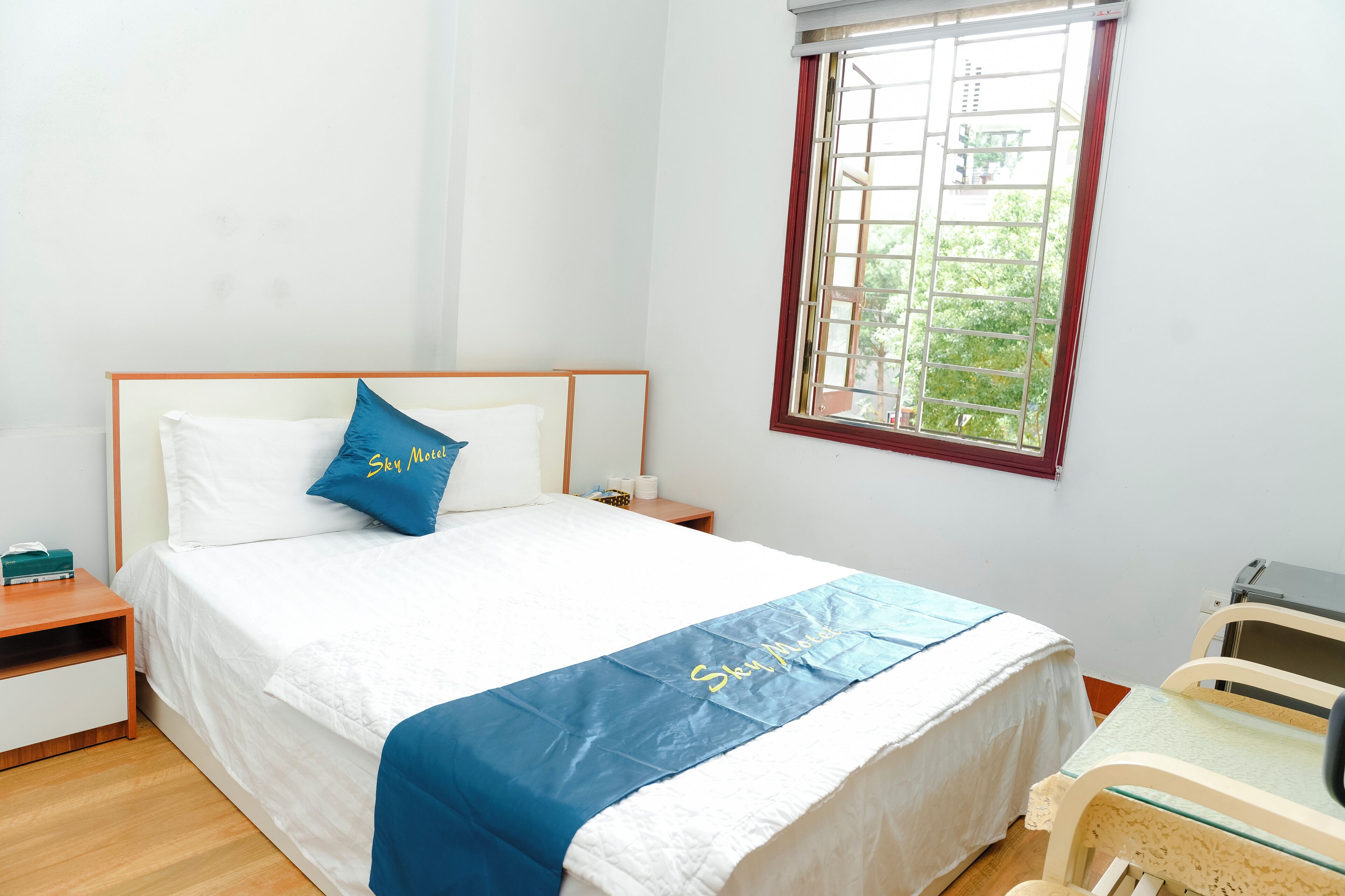 Ein ordentlich bezogenes Bett mit blauem Kopfkissen und weißer Decke, ein Nachttisch mit einem grünen Gegenstand, ein Fenster mit Blick auf Grünflächen und ein kleiner Tisch mit einem Stuhl.