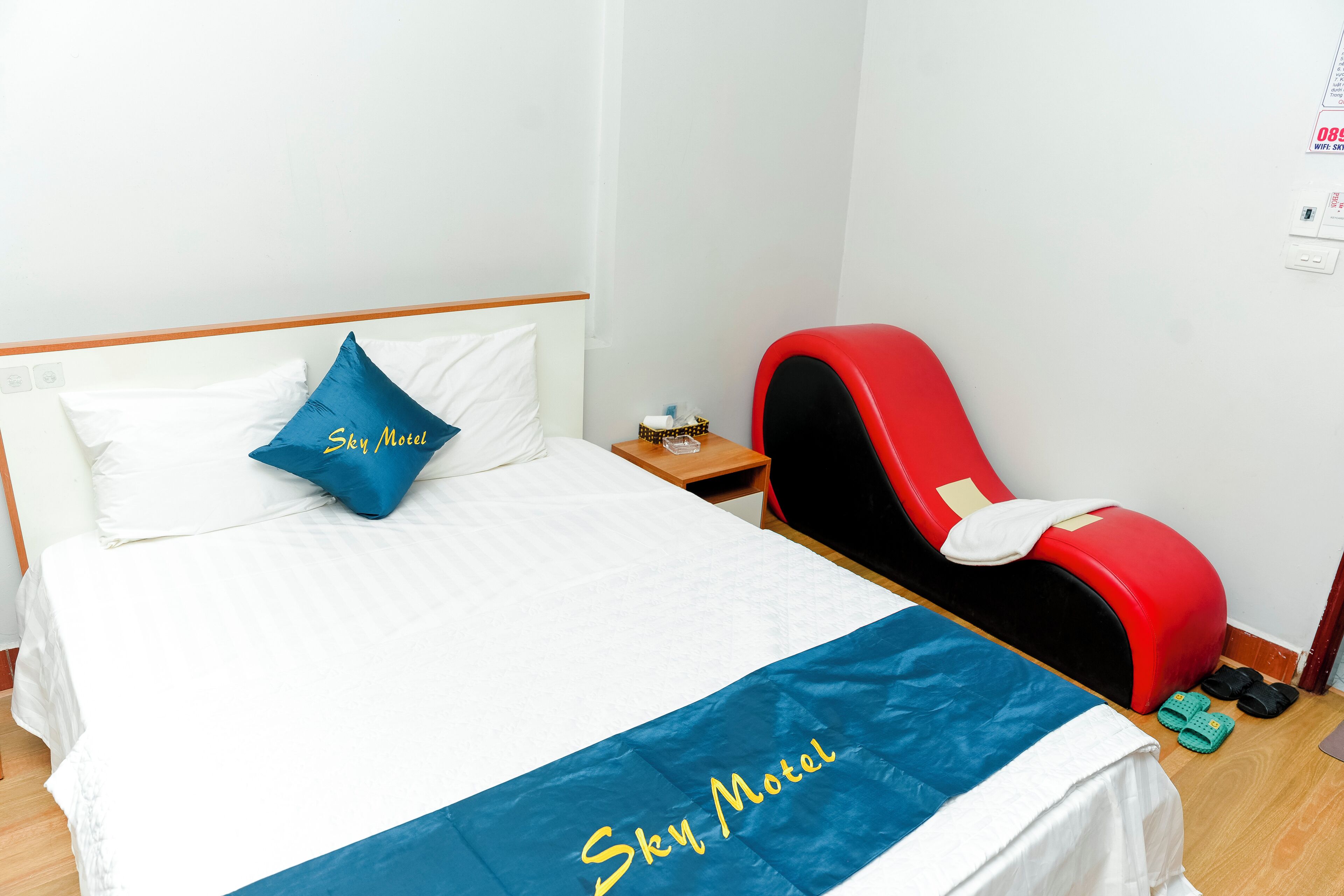 A hotel room with a bed, a red massage chair, slippers, and a bedside table with a tissue box.