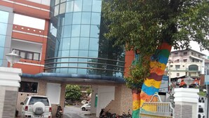 A modern building with a glass facade, a colorful vertical structure, and a gated entrance with parked vehicles. - Hotel Kurunji Meridian (Kodaikanal)
