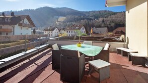 A terrace with wicker furniture, a table with a green tablecloth, and a view of a town and mountains.