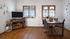 A room with a wooden ceiling, a TV on a wooden stand, a dining table with chairs, and windows with curtains.