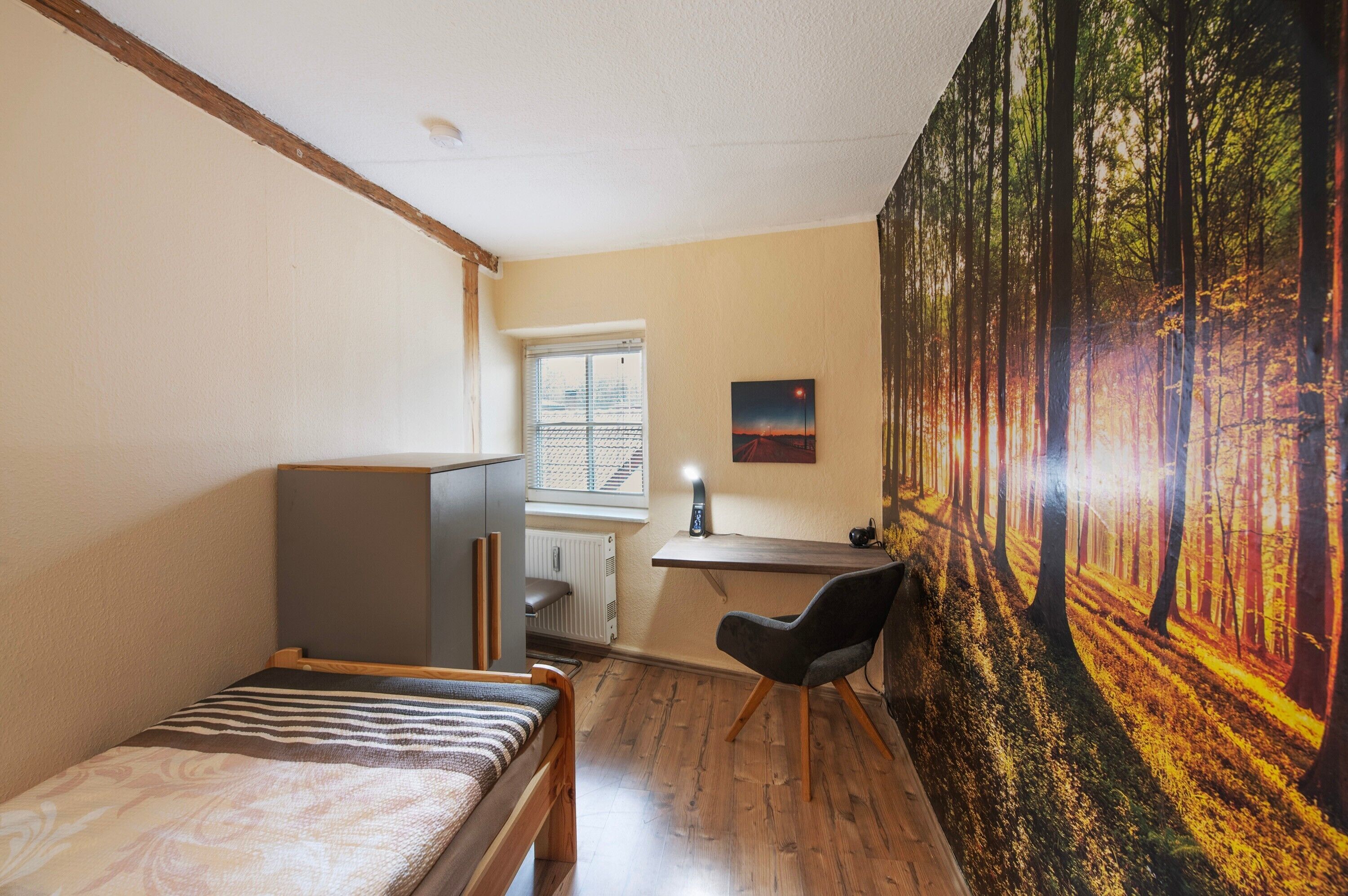 Una habitación con cama, escritorio, silla, radiador, ventana y un mural con motivos de bosque en la pared.