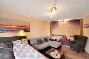 Una sala de estar moderna con un sofá modular, un sillón individual, una mesa de centro con un florero y arte en la pared que muestra un atardecer y pájaros.