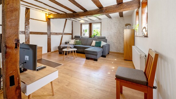 A room with exposed wooden beams, a grey sofa, a wooden chair, a small table with a computer, and a radiator. - Holiday Home 'Ferienhäuschen Kleine Auszeit' with Private Terrace, Private Garden and Wi-Fi (Seesbach)
