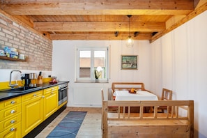 Une cuisine avec des meubles de couleur jaune, un plafond en bois, une fenêtre et un coin repas comprenant une table et un banc en bois.