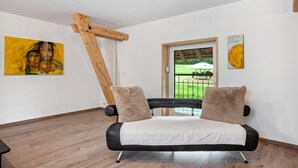 Ein modernes Zimmer mit einer schwarzen Couch, Holzbalken, einem Gemälde und Blick auf eine grüne Landschaft.