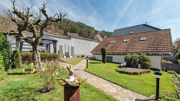 Une propriété résidentielle dotée d’un chemin en pierre, d’un jardin agrémenté de décorations colorées sur le thème de Pâques, et d’une maison avec un toit en tuiles et un espace extérieur couvert.