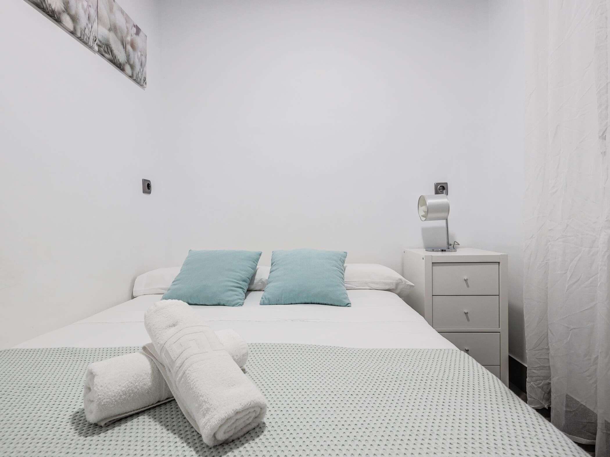 A minimalist hotel room with a bed, two pillows, a nightstand with a lamp, and a folded towel on the bed.