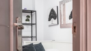 Un dormitorio moderno con una cama blanca, almohadas oscuras y un cabecero blanco. Hay una estantería con objetos decorativos y dos cuadros en la pared.