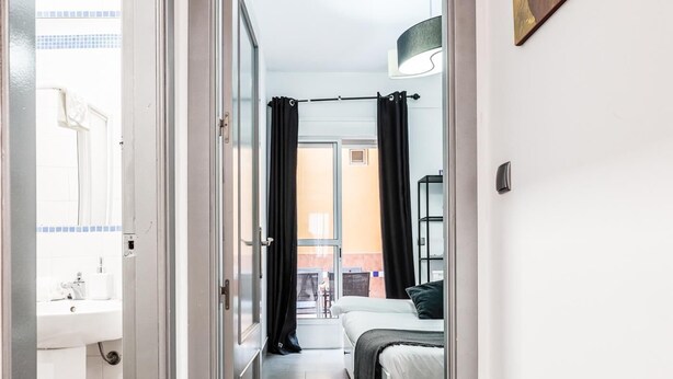 Un pasillo estrecho que conduce a un dormitorio con cama, ventana con cortinas y vistas al exterior.