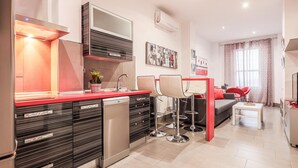 Una cocina moderna con cubierta roja, electrodomésticos de acero inoxidable y lavavajillas. Hay un comedor con mesa blanca y sillas rojas.