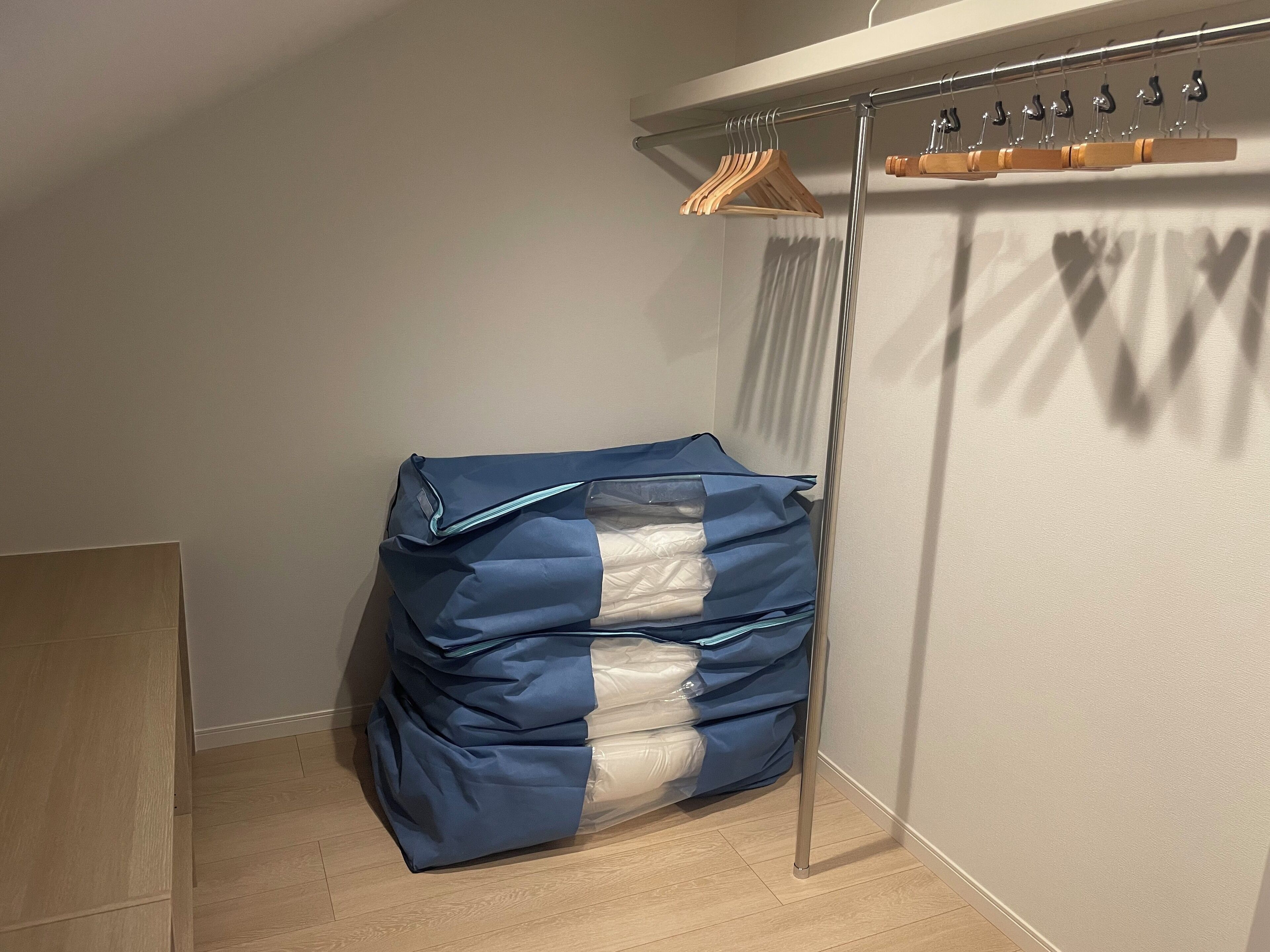 A corner of a room with a metal rack holding wooden hangers, a stack of folded clothes covered with blue and white plastic, and a wooden door frame.