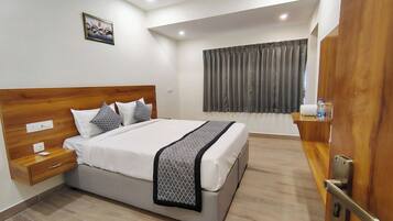 A modern hotel room with a wooden headboard, a large bed with white bedding, a bedside table with a remote control, and a view of the outside through a window with grey curtains.