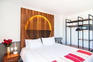 Een hotelkamer met een groot bed, een houten hoofdbord met een ronde lamp, een nachtkastje met een lamp en een metalen wandplank.
