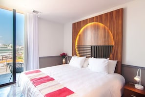 A hotel room with a large bed, a wooden headboard with a circular light, a nightstand with a lamp, and a view of the city through a large window.