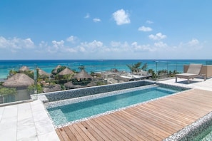 A pool area with a wooden deck, clear blue water, and a view of the ocean and thatched-roof structures.