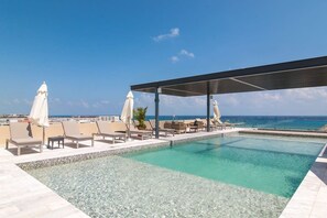 En pool på taget med liggestole og parasoller, med udsigt over en klar blå himmel og oceanet.