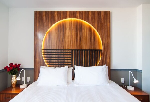 A hotel room with a large bed, two bedside tables, a wall-mounted circular light fixture, and a vase with red flowers.