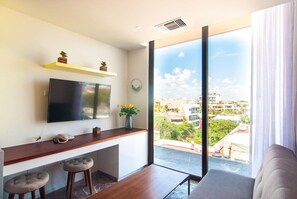 Een moderne woonkamer met een televisie, een houten bureau, twee comfortabele krukken, een kleine plant en een groot raam met uitzicht op de stad.