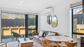 A modern interior with a sofa, dining table, chairs, and a large window with a view of a backyard. - Mt Roy Delight (Wānaka)