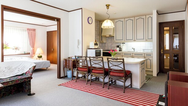 A compact hotel room with a bed, bedside table, lamp, and a small desk with a chair. A kitchen area is visible with a microwave, sink, and cabinets. - Eat and live like locals (Shinagawa City)