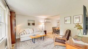 Una sala de estar con sofá, mesa de centro y sillón de cuero. Hay un comedor con mesa y sillas. Un televisor está montado en la pared.