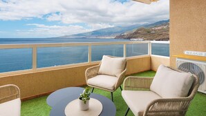 Un balcón con sillones de mimbre, una mesita con una planta y vistas al mar y colinas distantes.