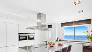 Una cocina moderna con armarios blancos, una isla central con taburetes y hornos integrados, y un amplio ventanal con vistas al mar.
