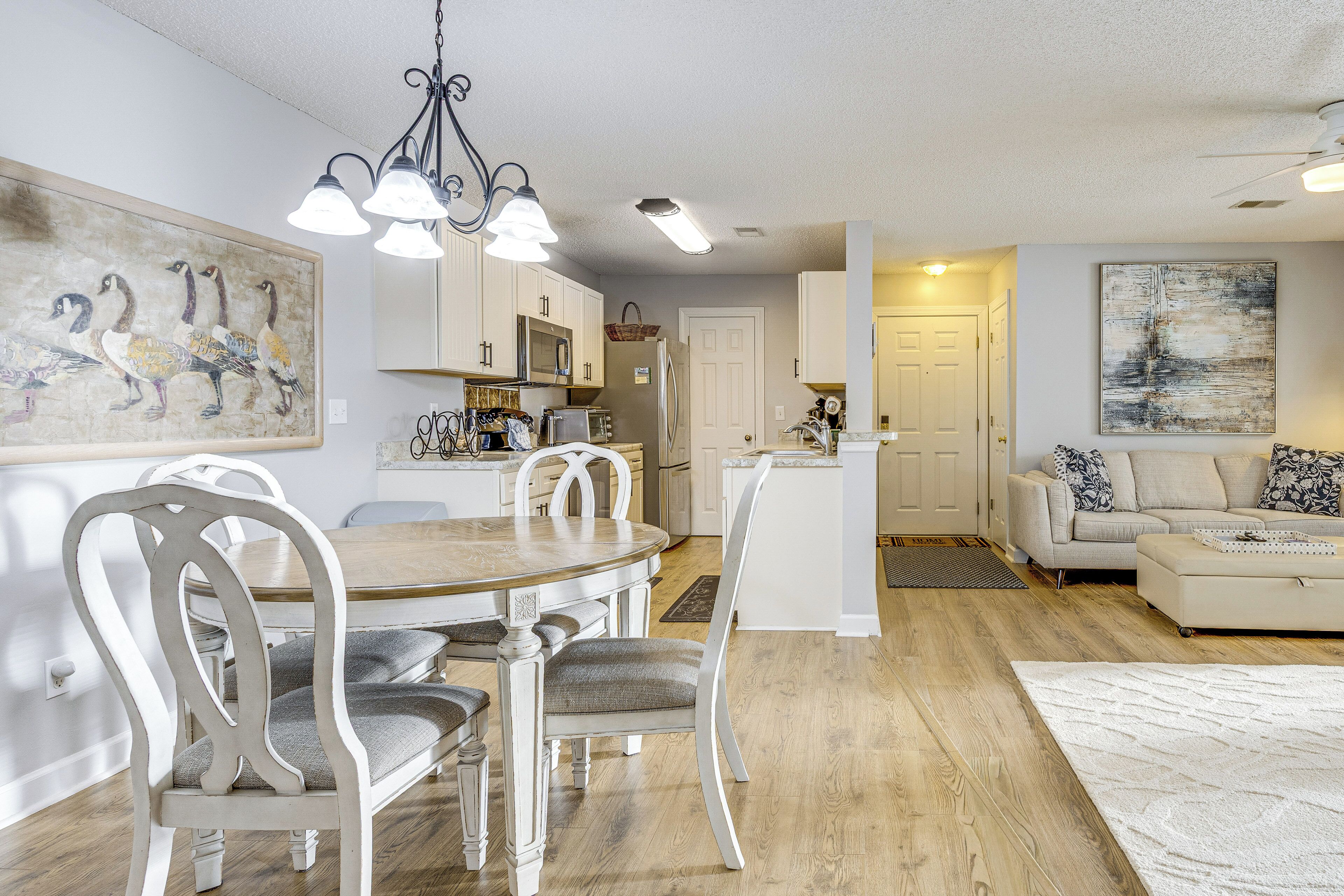 A modern kitchen with white cabinets, a dining table with chairs, a living area with a sofa, and artwork on the walls.