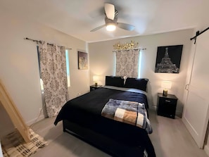 Un dormitorio con una cama grande, dos mesitas de noche con lámparas, cortinas estampadas, un ventilador de techo y obras de arte en las paredes.