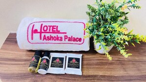 Ein Handtuch mit roten Stickereien, die "HOTEL Ashoka Palace" zeigen, drei kleine Flaschen mit dem Aufdruck "ASHOKA PALACE" und ein Topfpflanzen mit gelben Blüten.