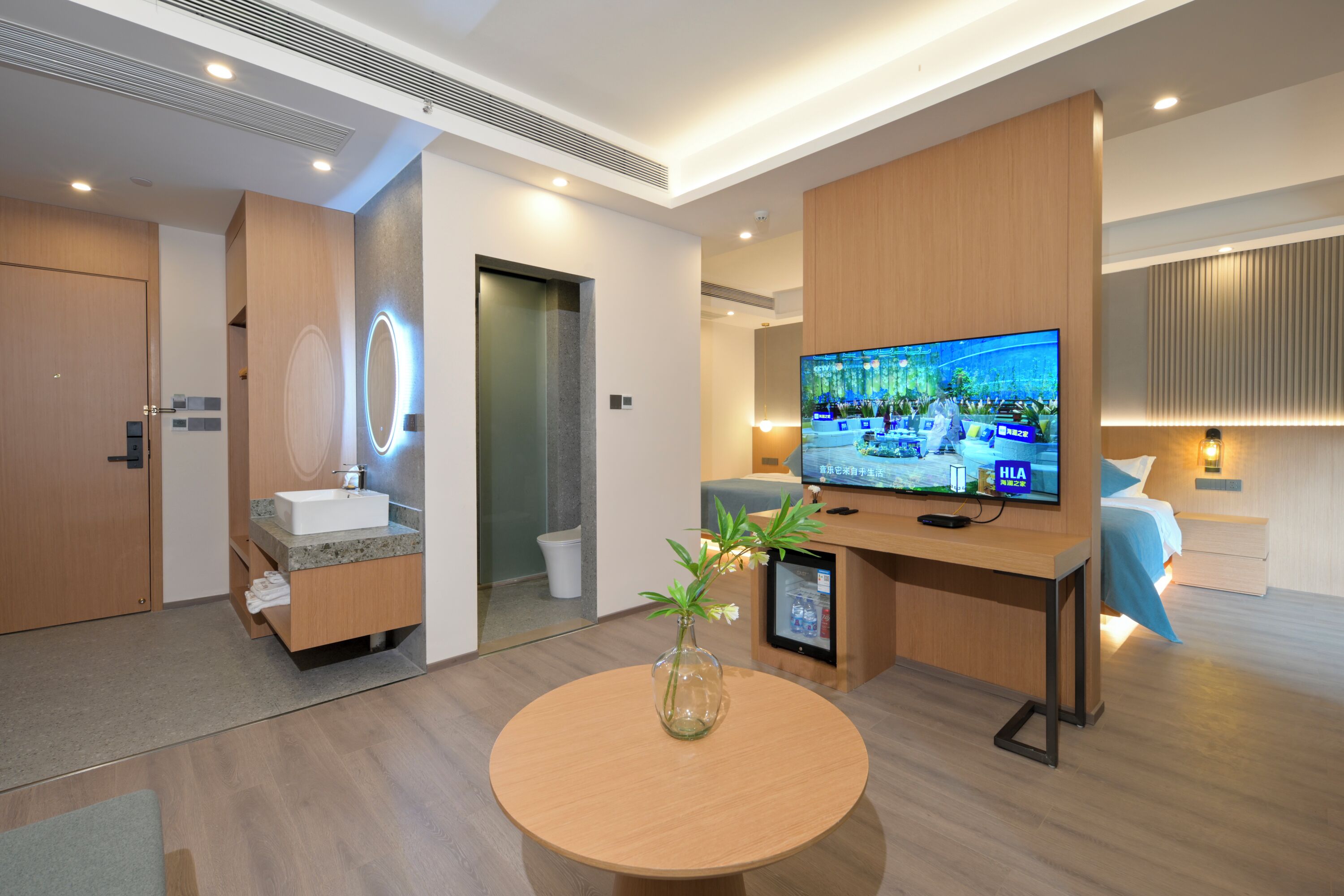 Una habitación de hotel moderna con un escritorio de madera, un televisor, una mesa redonda con un florero y un baño con lavabo y espejo.