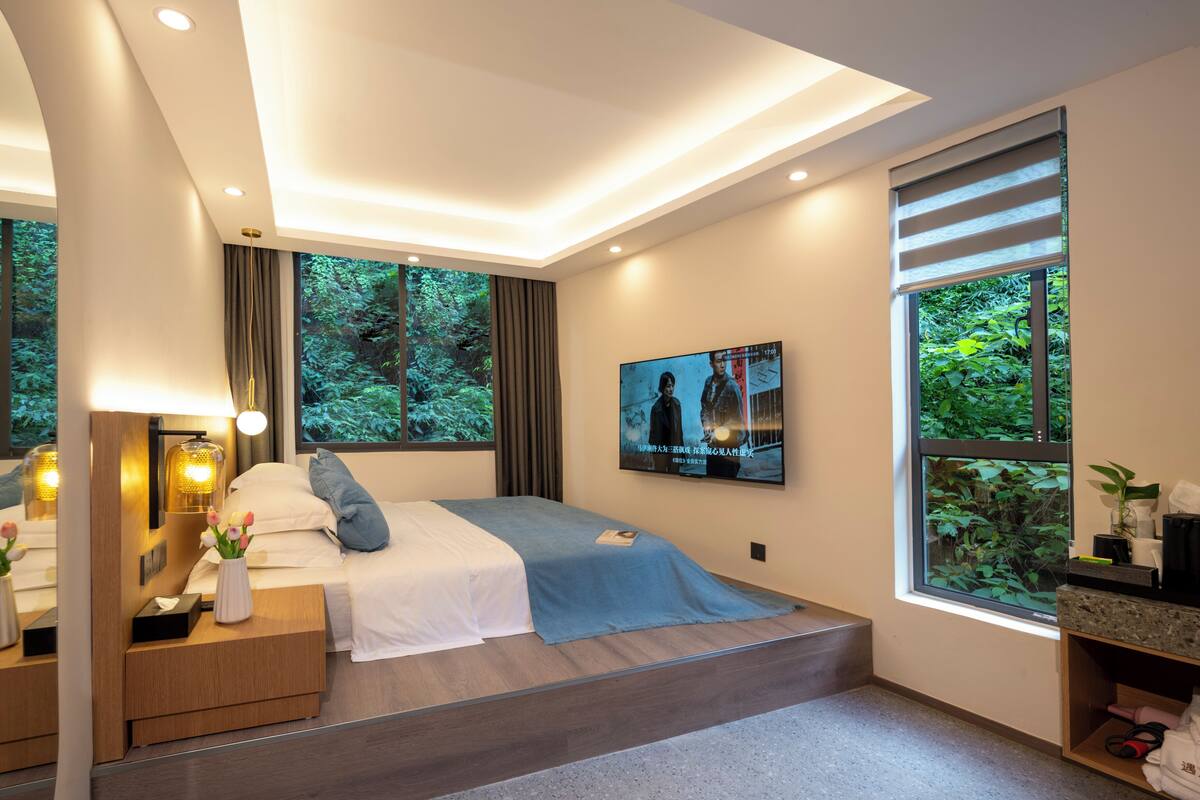 Quarto moderno com cama, mesas de cabeceira, televisão e janela com vista para vegetação.